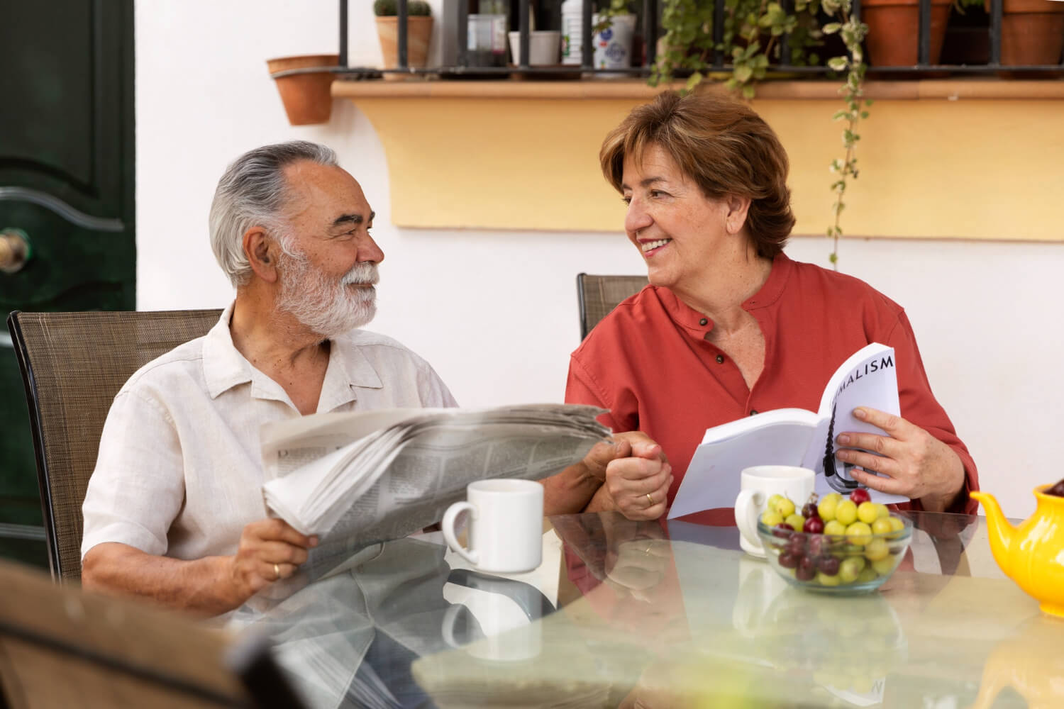 Happy senior couple enjoying a relaxed morning together with coffee, reading a newspaper and a book outdoors.